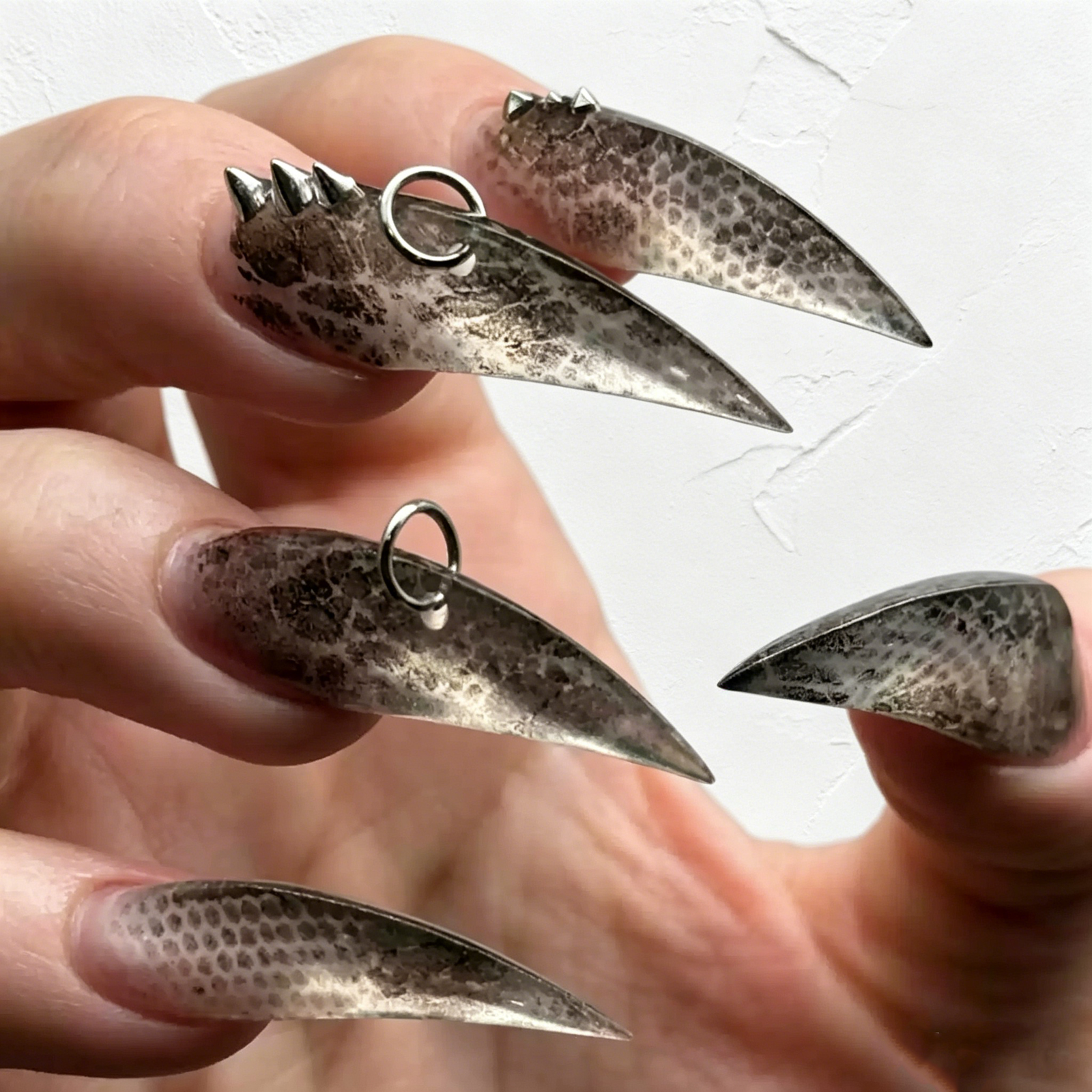 Close-up hand shot smoky black speckled stiletto press on nails with detachable metal ring charms, goth punk sharp claw fake nails edgy dark manicure