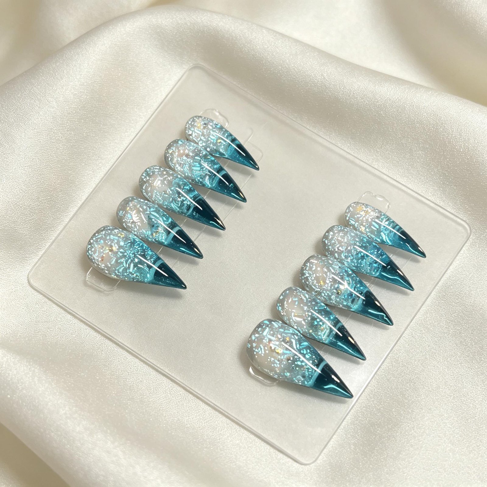 Shimmery aqua blue foil stiletto press on nails flat lay on soft white satin fabric